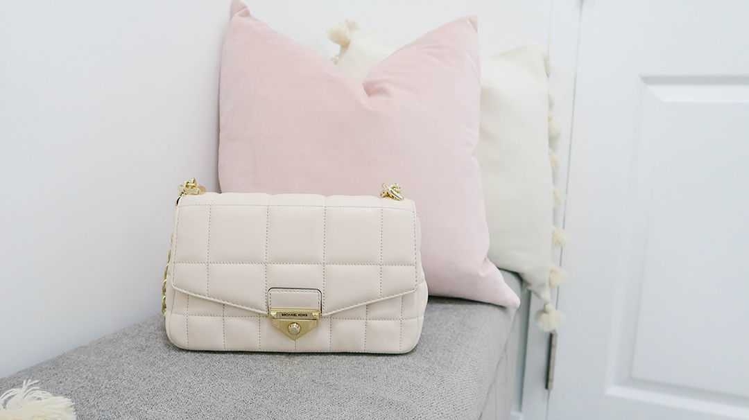 Michael Kors Soho Purse sits on a gray bench with pink throw pillows