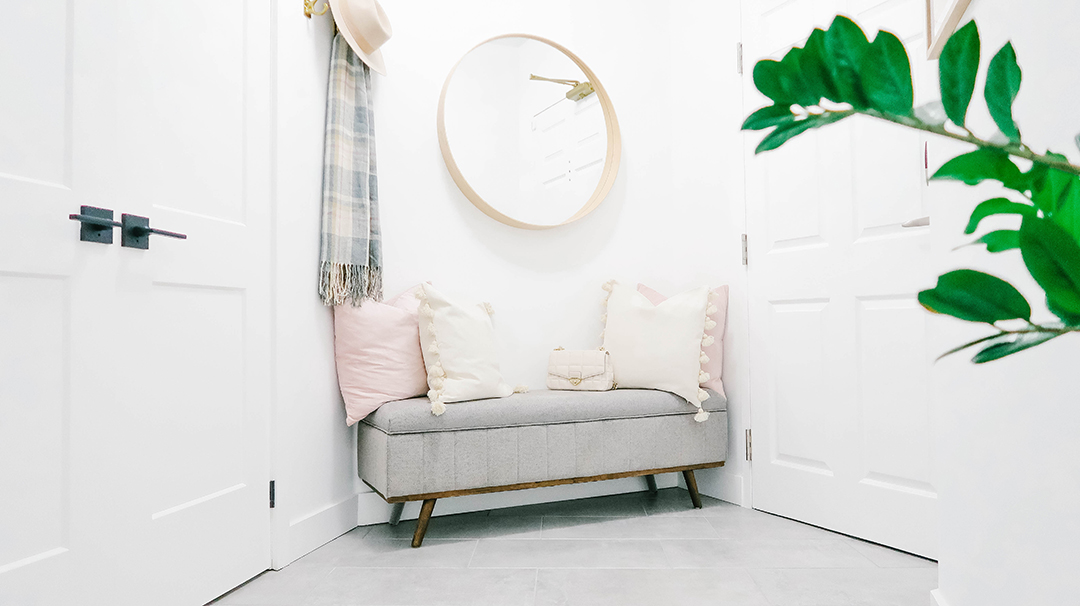 A gray bench sits with cozy pillows and round mirror above and hook to the side with a hat and scaf